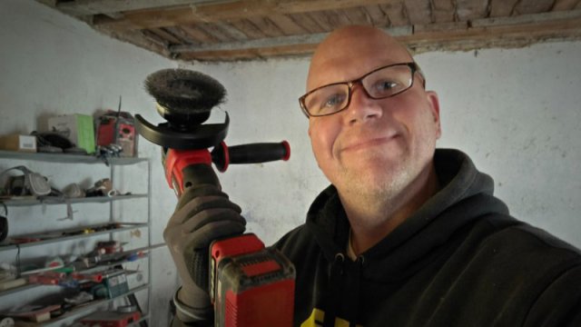 Ein Mann mit Brille und schwarzem Kapuzenpullover h&auml;lt l&auml;chelnd einen Akku-Winkelschleifer mit Drahtb&uuml;rstenaufsatz.