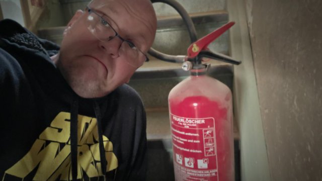 Ein Mann mit Star-Wars-Kapuzenpullover neben einem roten ABC-Pulver-Feuerl&ouml;scher auf einer Baustellentreppe.