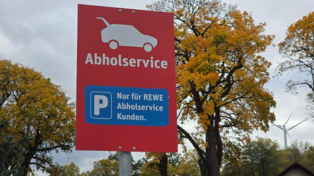 Rotes REWE Abholservice Schild auf einem Parkplatz vor herbstlichen B&auml;umen